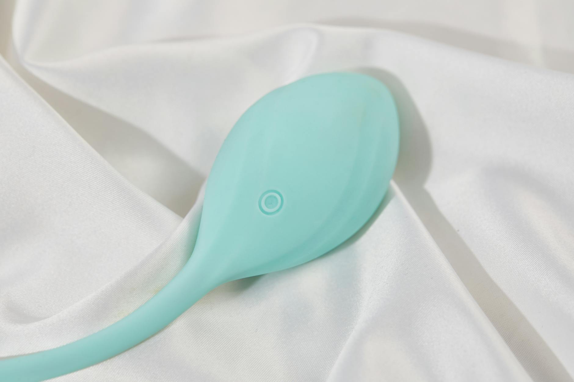 A teal clitoral vibrator rests on smooth white silk fabric, symbolizing pleasure and intimacy