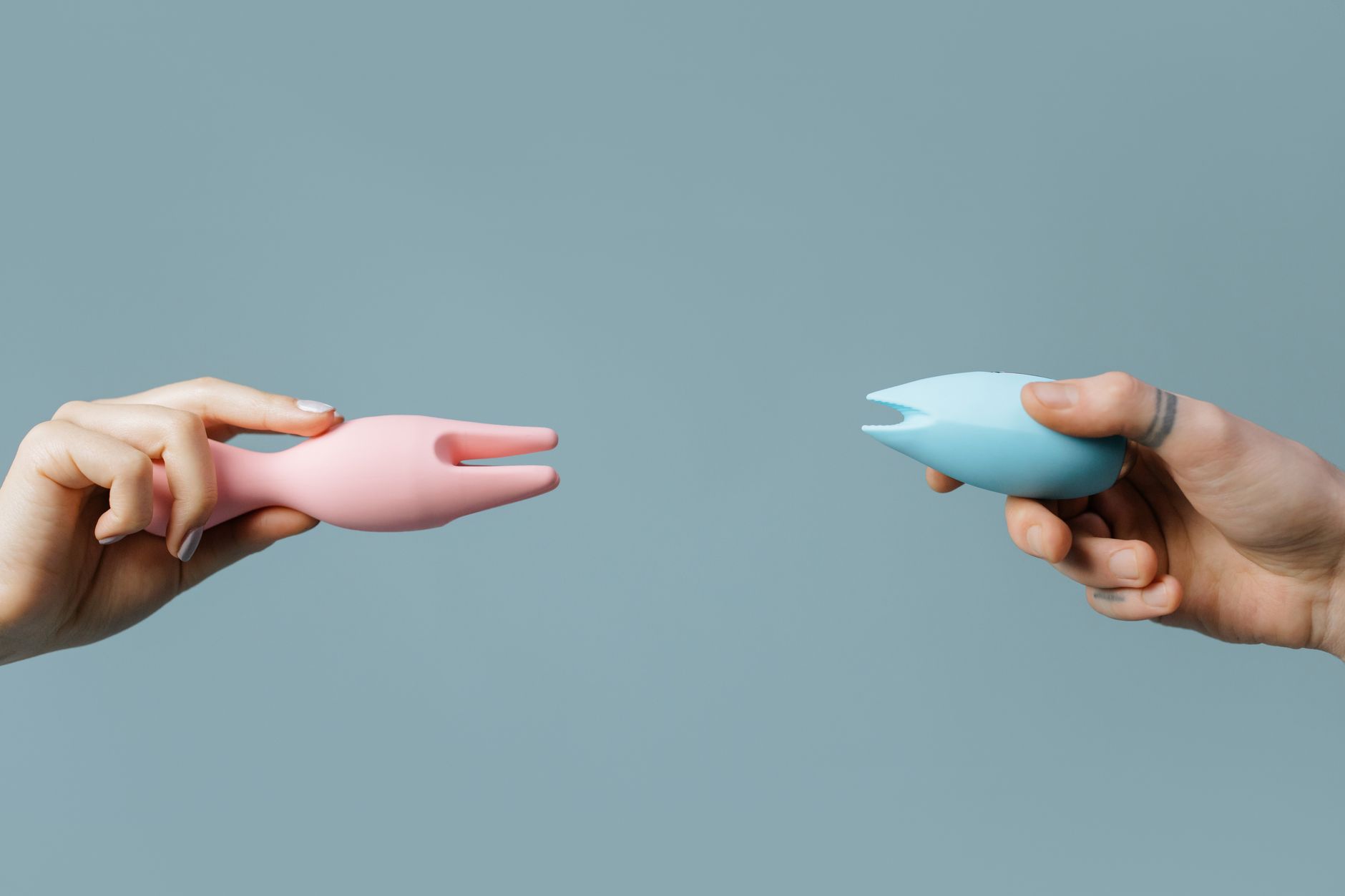 Two hands holding pink and blue silicone vibrators on a pastel background